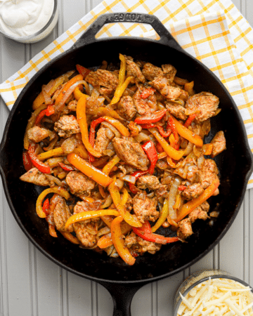 A cast iron skillet, filled with chicken fajita ingredients: chicken breast, sliced peppers, and onions. The skillet rests on a tea towel, and adjacent to a bowl of sour cream, as well as a bowl of shredded cheese.