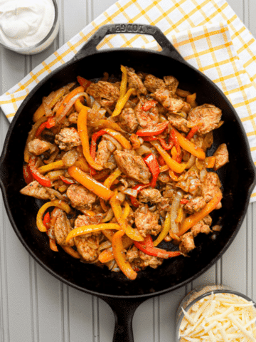 A cast iron skillet, filled with chicken fajita ingredients: chicken breast, sliced peppers, and onions. The skillet rests on a tea towel, and adjacent to a bowl of sour cream, as well as a bowl of shredded cheese.