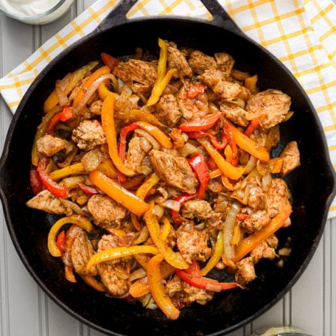 A cast iron skillet, filled with chicken fajita ingredients: chicken breast, sliced peppers, and onions. The skillet rests on a tea towel, and adjacent to a bowl of sour cream, as well as a bowl of shredded cheese.