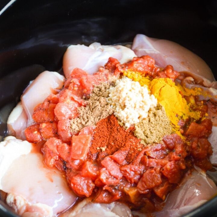 A slow cooker filled with raw chicken, spices, and tomato.