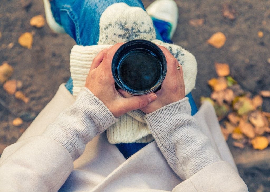 A woman is holding a coffee cup, wearing a sweater and jeans. Fallen leaves are in the background.