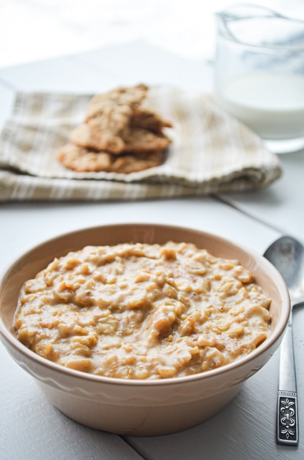 A bowl of peanut butter cookie oatmeal, with peanut butter cookies and a cruet of milk in the background.