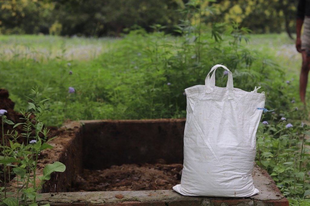 A large feed bag, filled with compost, sits on the ledge of a stone-edged compost pile. Purchasing and ordering compost is a great way to prepare for gardening season.