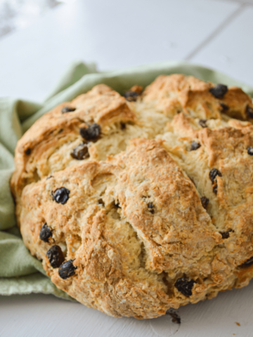 A loaf of Irish Soda bread lays on top of a green cloth napkin. A large cross is cut in the center and it is dotted with raisins.