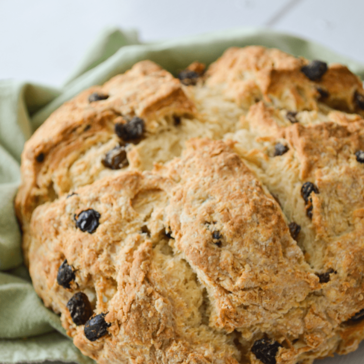 A loaf of Irish Soda bread lays on top of a green cloth napkin. A large cross is cut in the center and it is dotted with raisins.