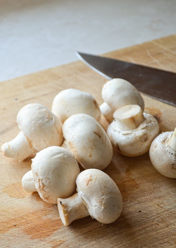 A wood cutting board with button mushrooms resting on top. A large chefs knife to the right.