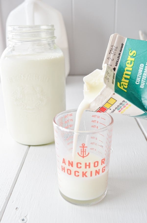 Store bought buttermilk is being poured into a small glass measuring cup. A jar of whole milk is to the left.