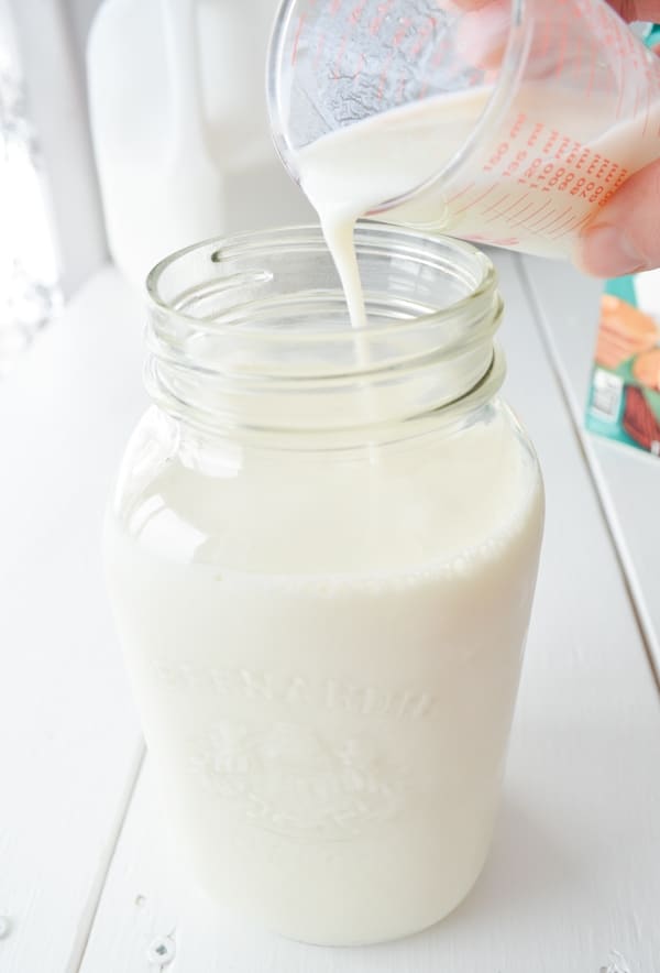 A small measuring cup is pouring cultured buttermilk into a glass jar of milk.