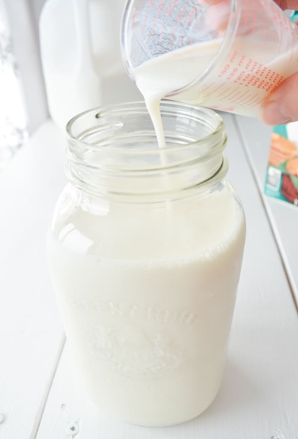 A small measuring cup is pouring cultured buttermilk into a glass jar of milk.