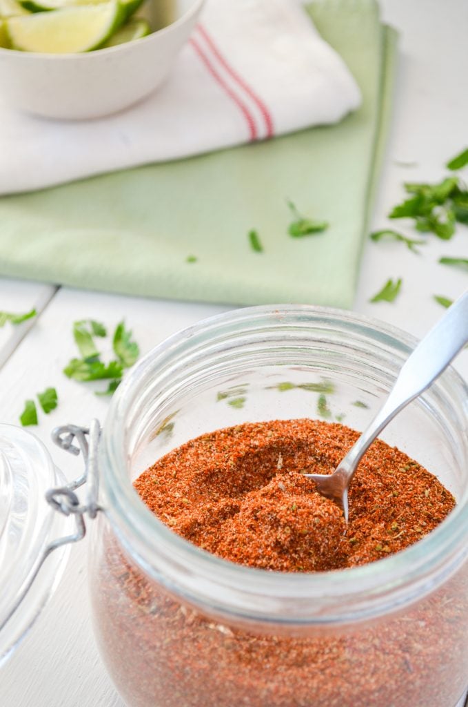 A jar of chicken taco seasoning rests on a white table. Cilantro is sprinkled in the background.