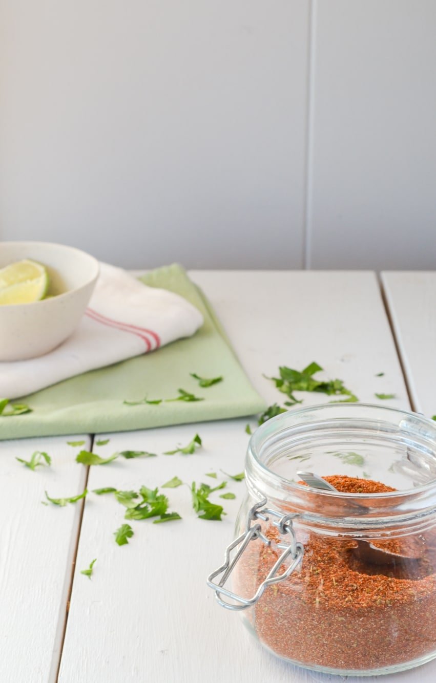 A white table with a jar of a spice mixture, cilantro sprinkled in the background, and two cloth napkins with a bowl of lie wedges.