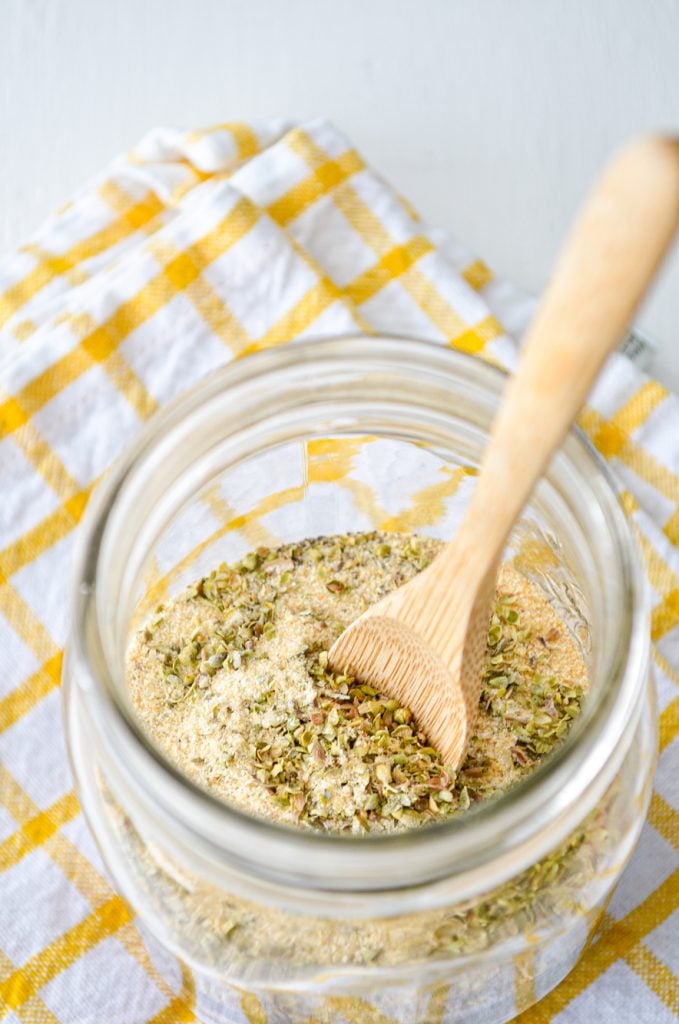 A jar of spices with a small wooden spoon rests on a white and yellow cloth towel.