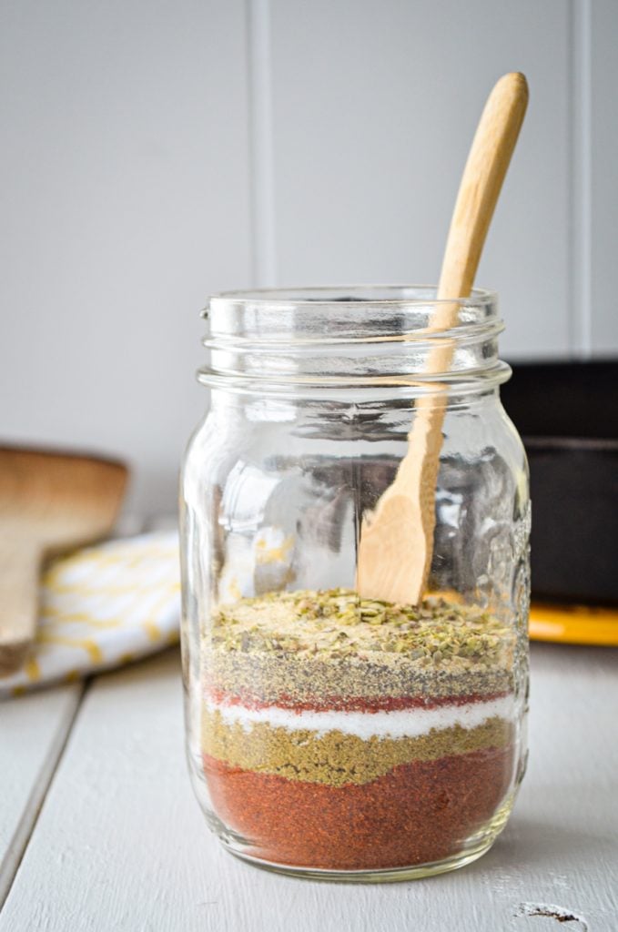 A jar of layered spices with a small wooden spoon inside. A cast iron skillet, wooden spatula, and tea towel are in the background.