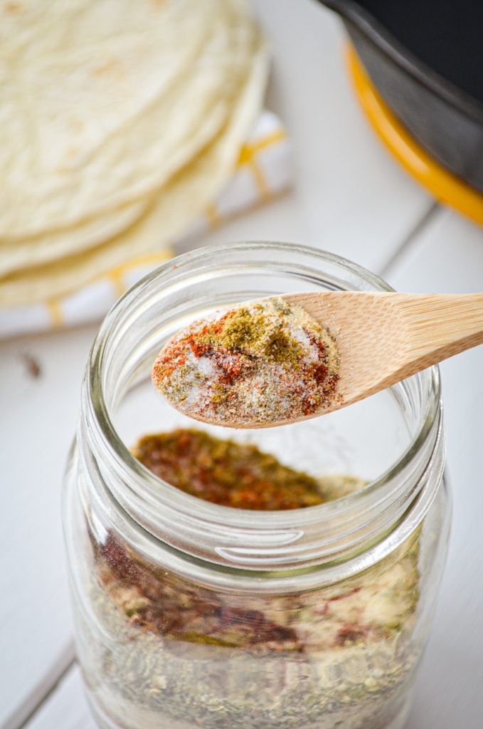 A jar of blended spices is being spooned out of a pint sized jar. A stack of tortillas in the background.