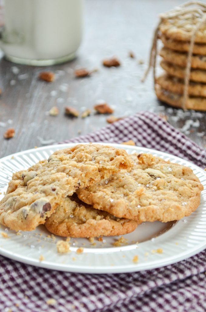 A plate of cookies on a cloth napkin. A glass of milk and stack of cookies in the background.