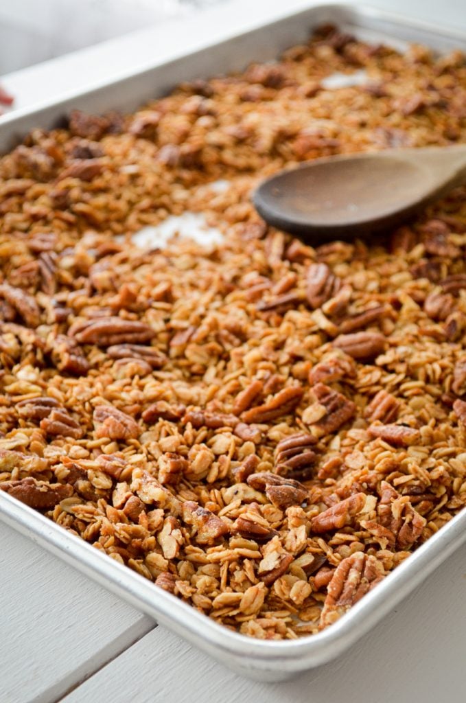 A sheet pan, filled with butter pecan granola, and a wooden stirring up the granola.
