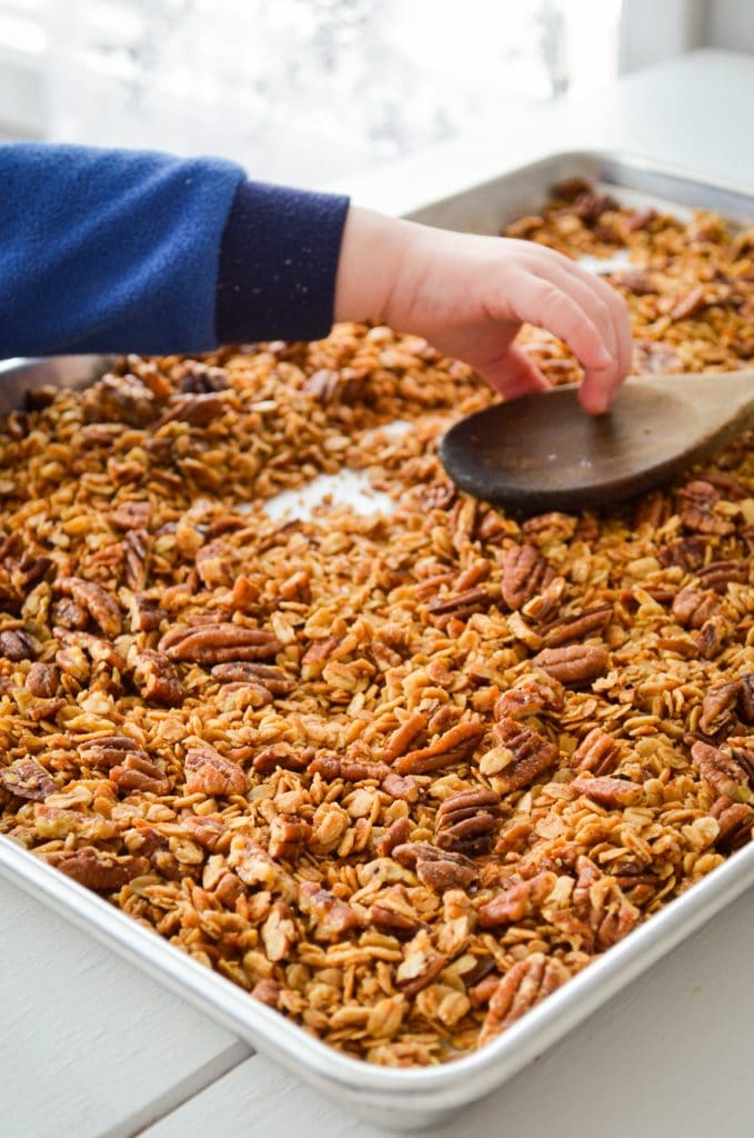 A toddler hand reaches in to grab a wooden spoon from a sheet pan of butter pecan granola.