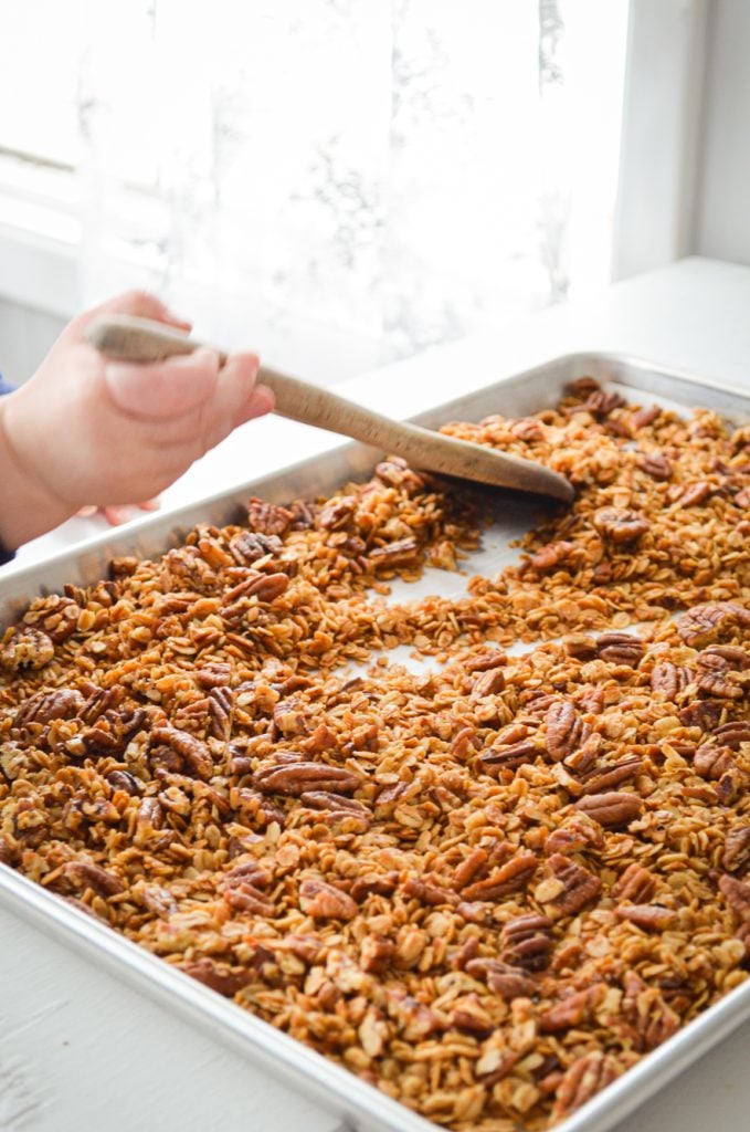 A toddler hand, using a wooden spoon to stir up the granola.