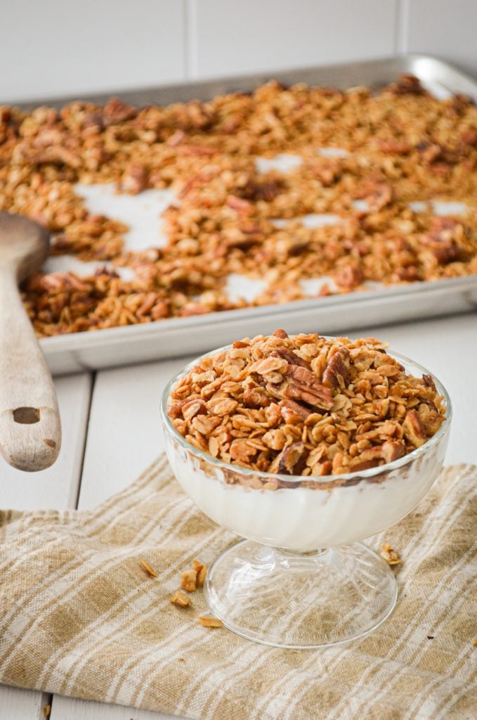 A yogurt parfair, topped with granola. A sheet pan of granola in the background, with a wooden spoon.