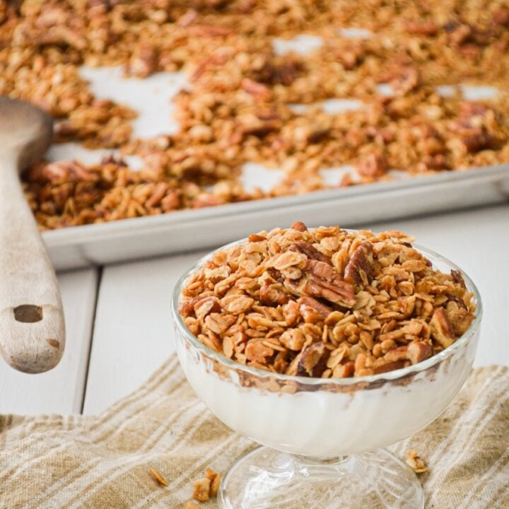 A yogurt parfair, topped with granola. A sheet pan of granola in the background, with a wooden spoon.