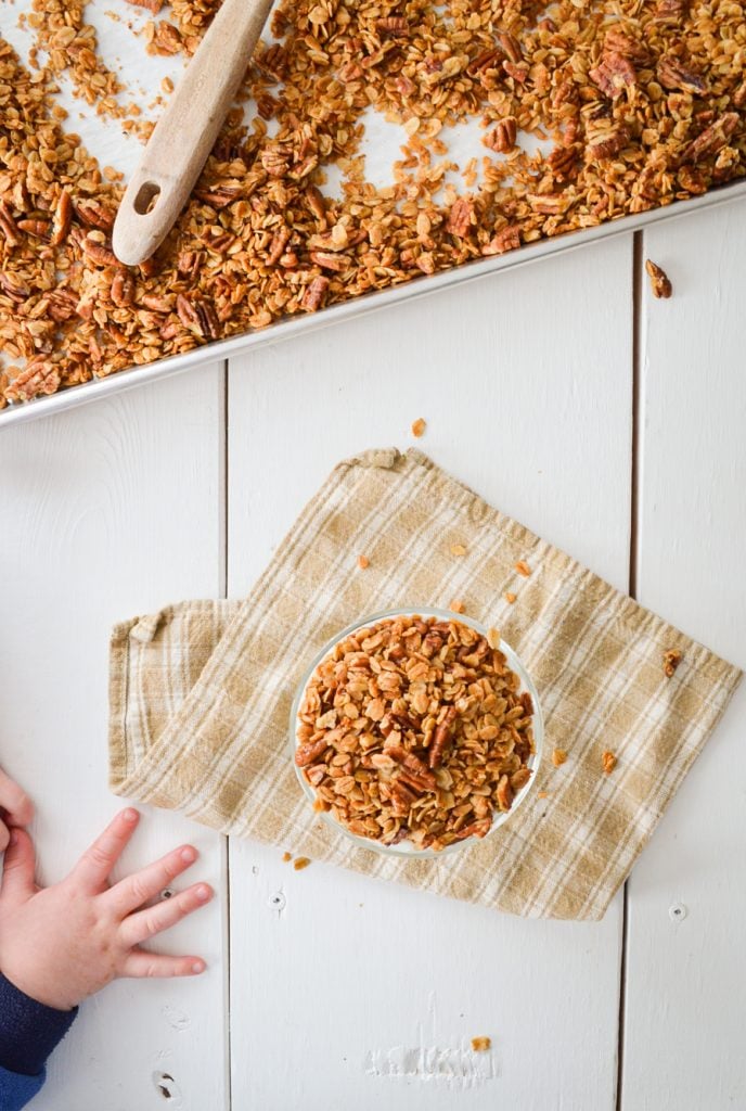 A birds eye view of a sheet pan of granola, a yogurt parfait, and a toddler hand reaching in.