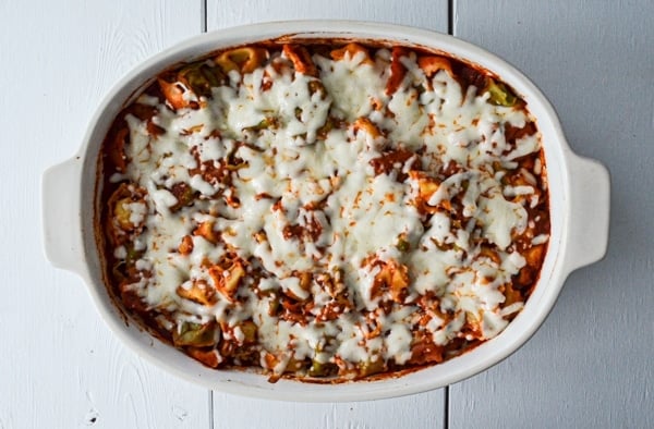 A casserole of tortellini, tomato sauce, and cheese is baked in a 9X13 pan on a white wood background.