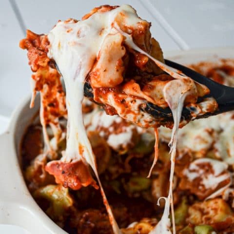 A baking dish of cheesy tortellini bake, with a portion being pulled out. Big strings of cheese connect the serving to the casserole.