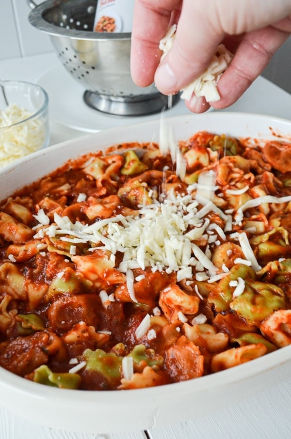 A hand sprinkles cheese over a baking dish filled with tortellini and pasta sauce.