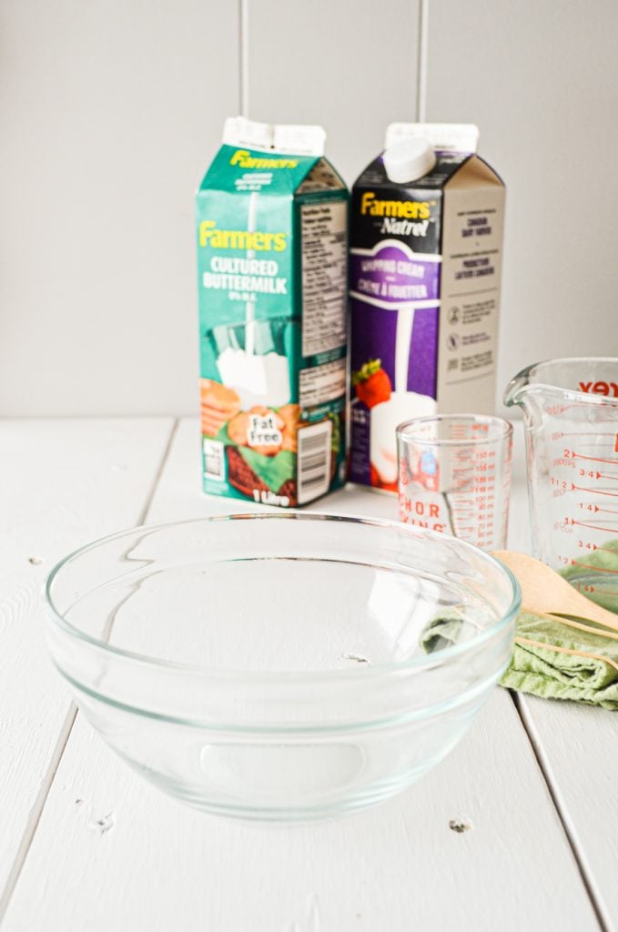 A glass bowl rests on a table, with a carton of heavy cream and a carton of buttermilk in the background.