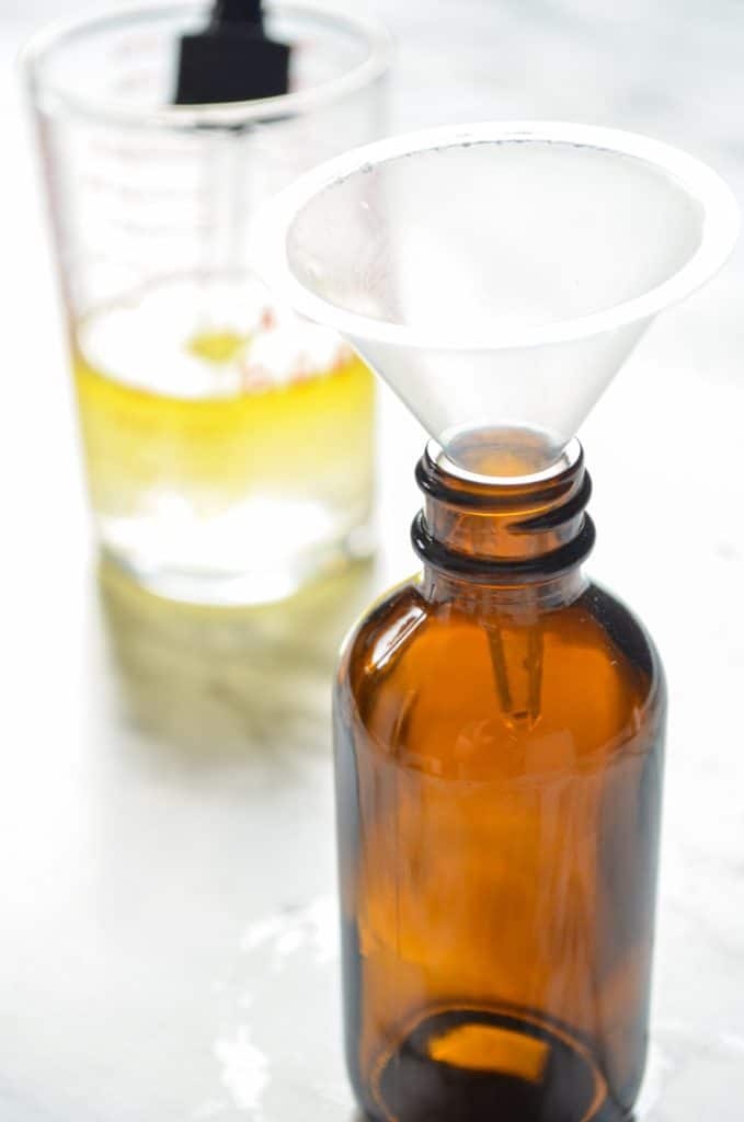 A small amber jar fitted with a funnel. A small measuring cup in the background with DIY facial serum in it.