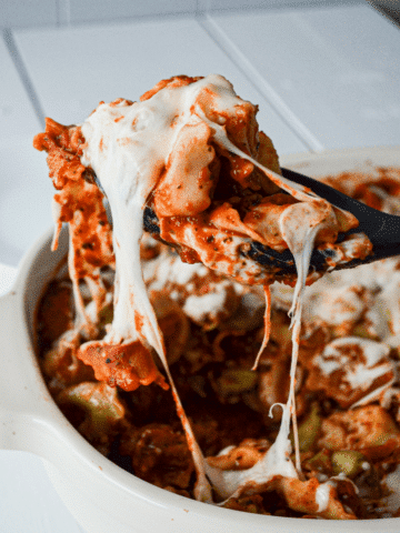 A large spoon removes a serving of cheesy tortellini casserole from a baking dish.