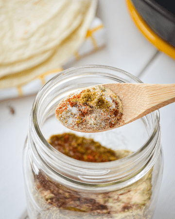 A jar of homemade taco seasoning, with a small wooden spoon lifting the contents. A stack of tortillas in the background.
