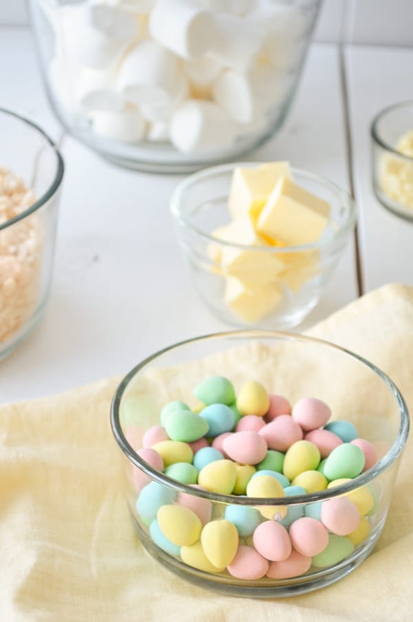 A glass bowl of mini eggs in the bottom right corner. A bowl of cubed butter, rice krispies, and marshmallows can be seen in the background.