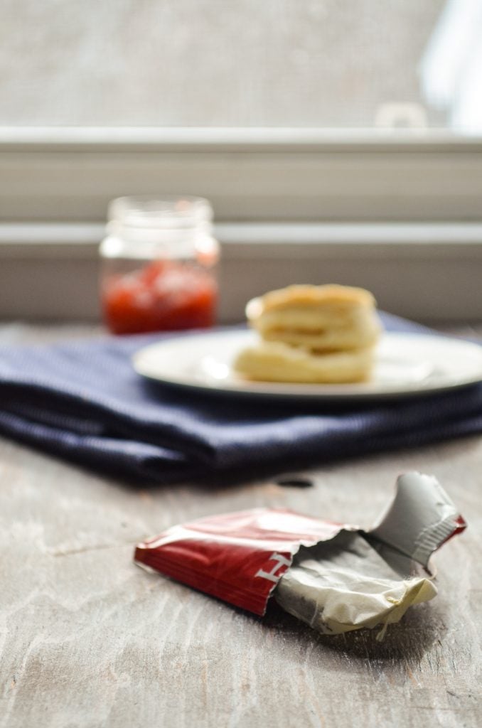 A chai tea bag, with a biscuit and a small jar of strawberry preserves in the background.