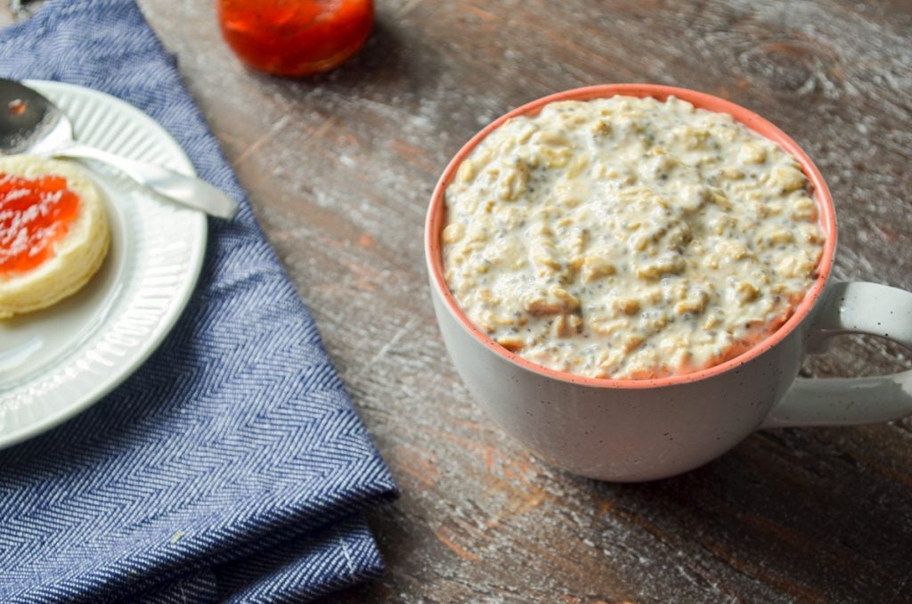 A mug of overnight oats, with a biscuit and some strawberry preserves in the background.