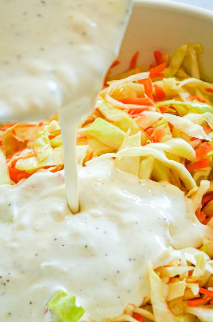 A close up of buttermilk coleslaw dressing being poured on top of shredded cabbage and carrots.