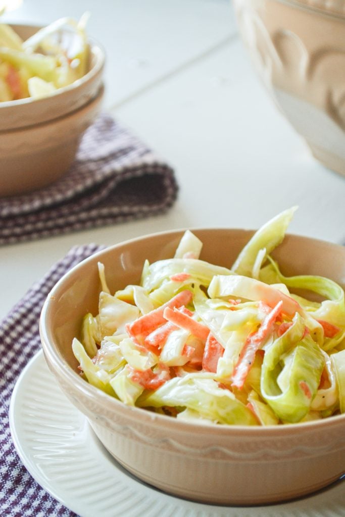 A close up of a bowl of buttermilk coleslaw, resting on a purple napkin. A second bowl in the back left, and the serving bowl in the back right.