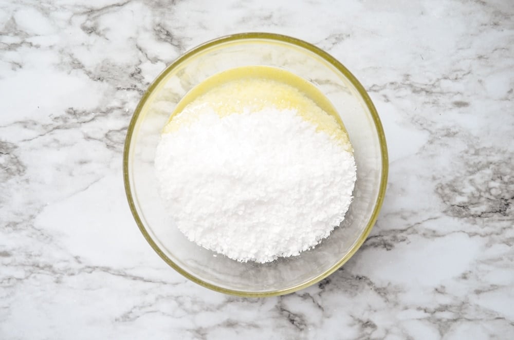 An overhead view of the process of making a DIY facial salt scrub. Coarse salt, coconut oil, and jojoba oil are all combined in a small bowl.