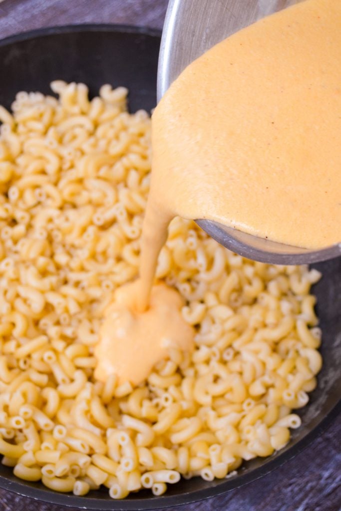 A homemade cheese sauce being poured over cooked elbow macaroni.