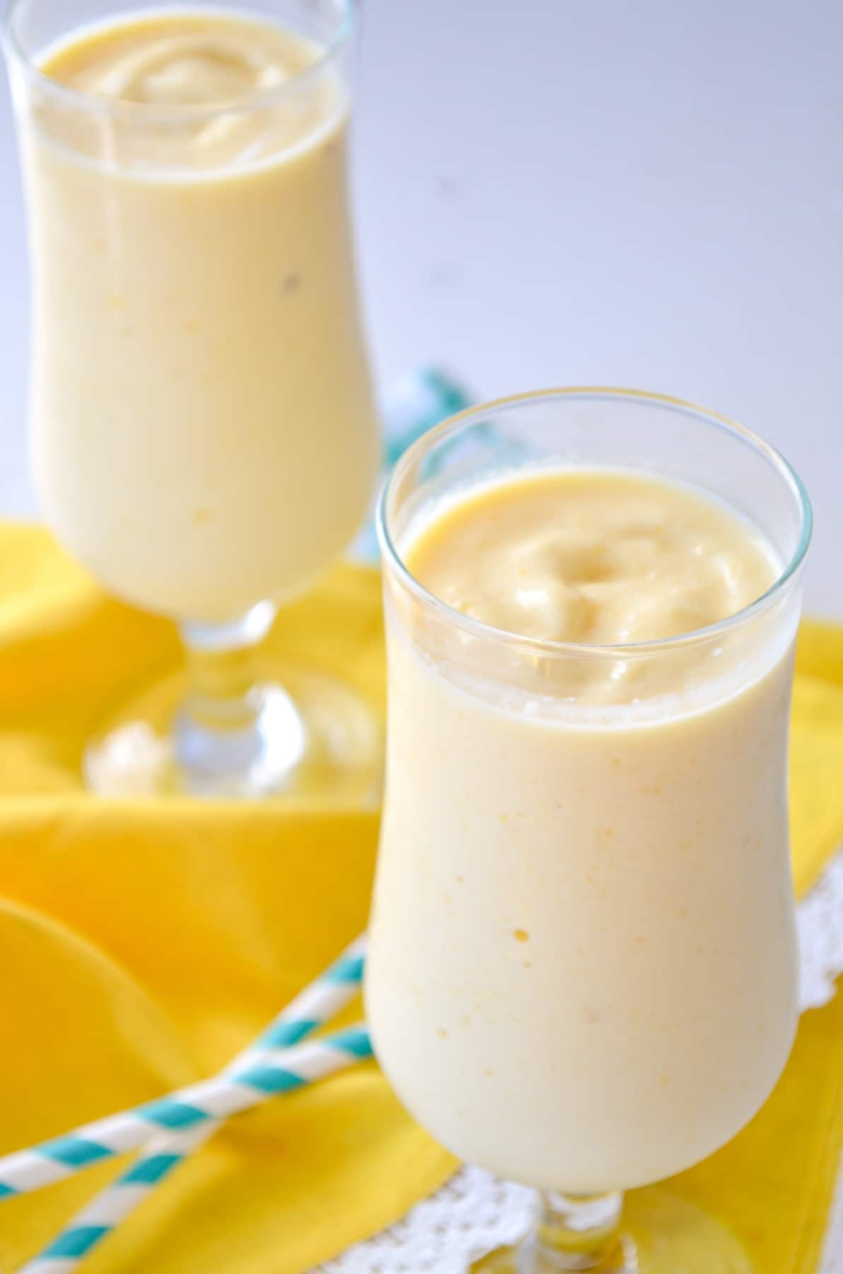 Two glasses of mango pineapple smoothie on a yellow napkin with straws on top.