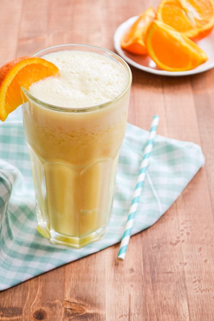 A glass of copycat orange julius, a plate of sliced oranges in the background.