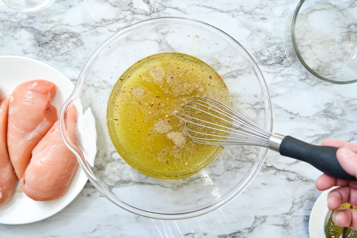 An overhead shot of a bowl of pickle juice, whisked together with various seasonings. A plate of raw chicken breasts to the left hand side.