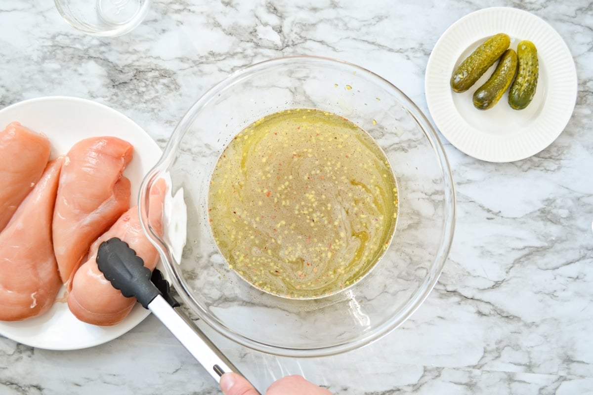 An overhead shot of a bowl of pickle brine. To the left, a pair of tongs is picking up a chicken breast from a plate.