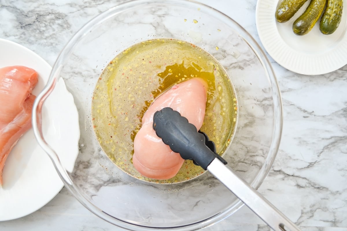 An overhead shot of a pair of tongs placing chicken breasts into a brine of pickle juice.