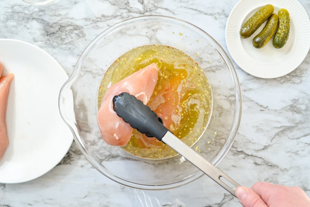 An overhead shot of a pair of tongs lifting chicken breasts into a bowl of pickle juice brine.