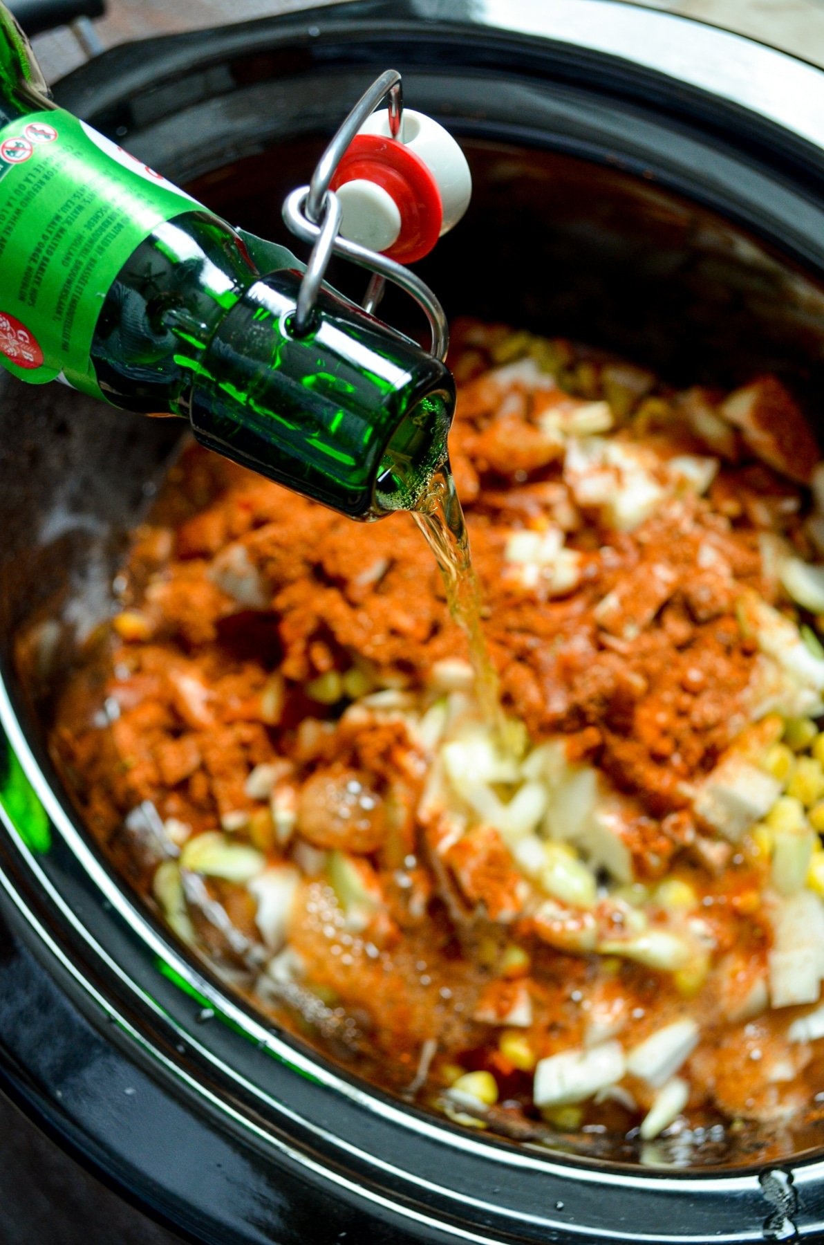 A bottle of beer being poured into a slow cooker full of chicken taco soup.