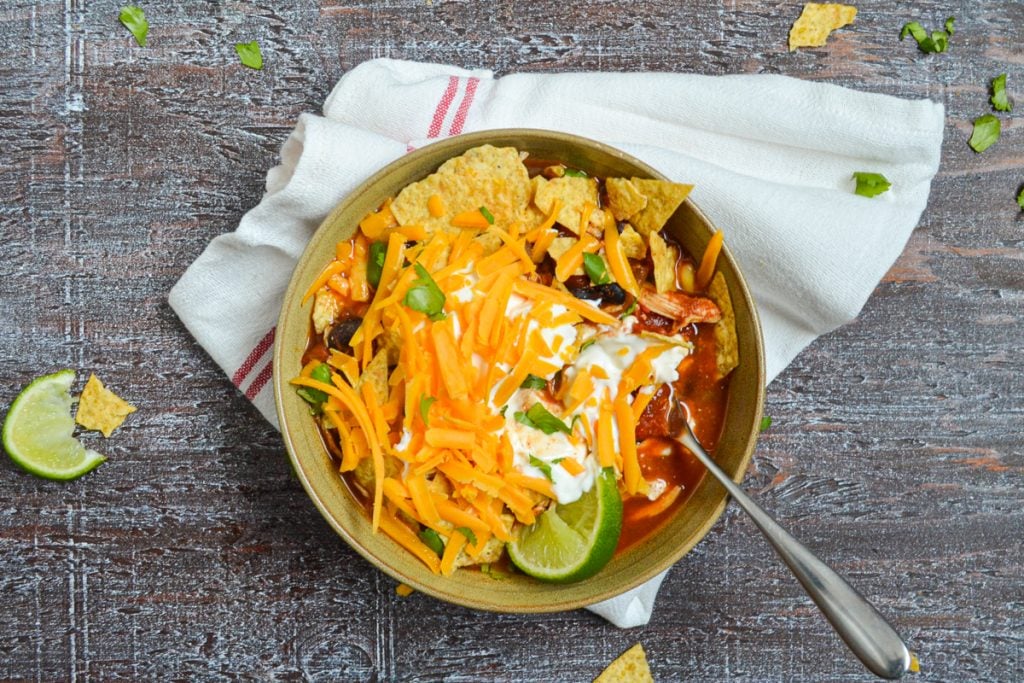 A bowl of chicken taco soup, topped with shredded cheese, sour cream, crushed tortilla chips, and lime wedges.