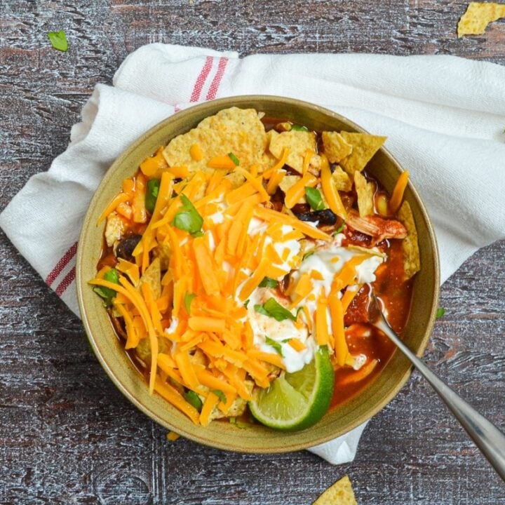 A bowl of chicken taco soup, topped with shredded cheese, sour cream, crushed tortilla chips, and lime wedges.