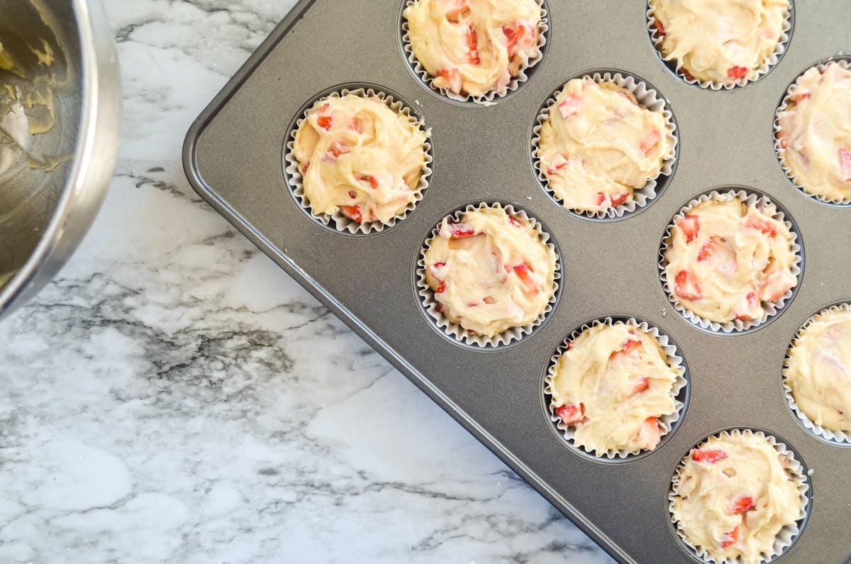 Strawberry buttermilk muffins inside a muffin tin, with a bowl of batter off to the left.