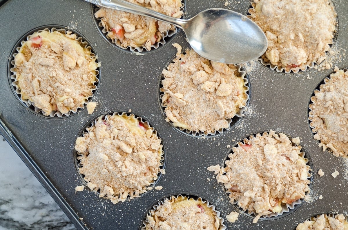 Muffins in a muffin tin, with a streusel topping. A spoon resting on the muffin tin. 